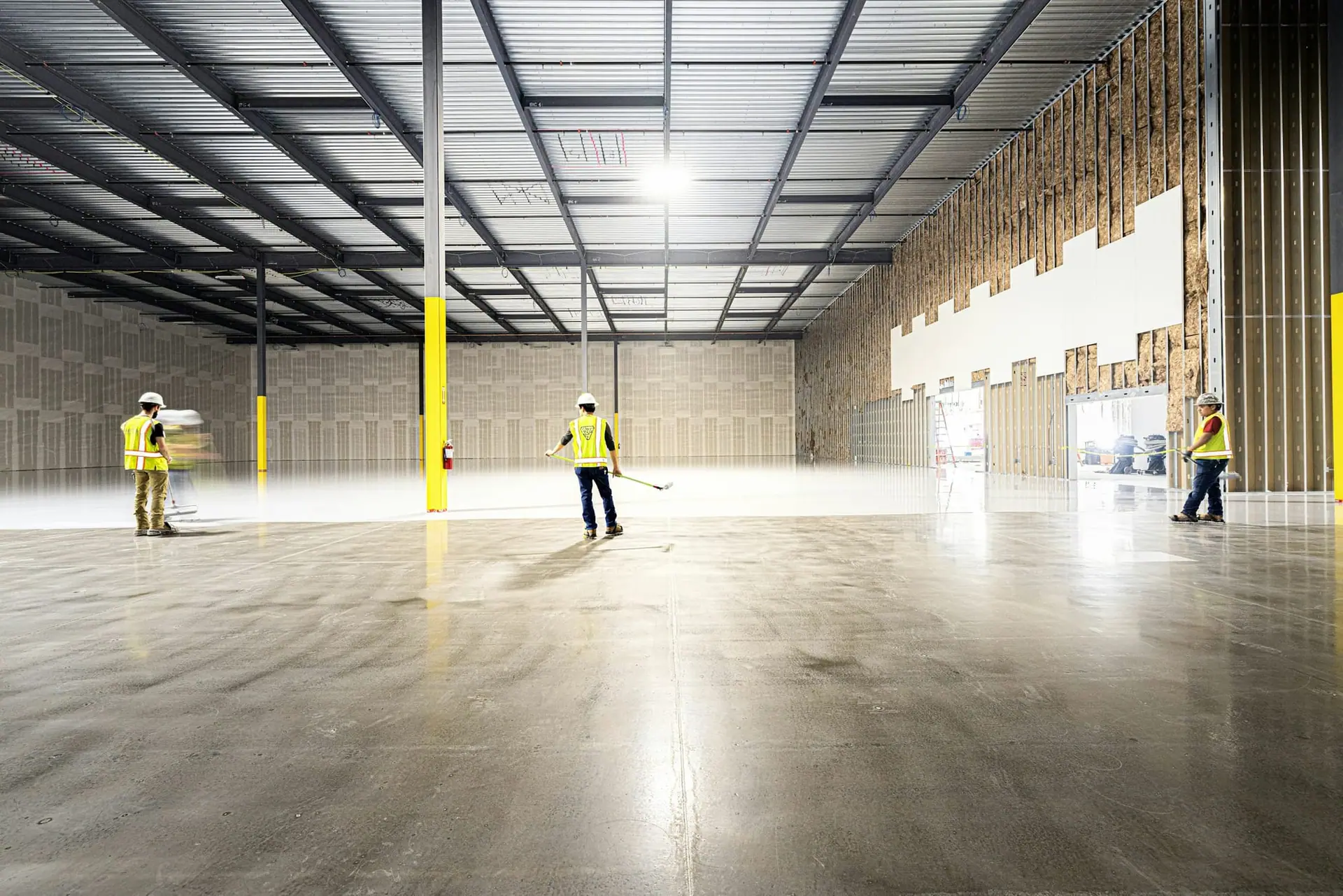 Large empty warehouse interior with steel roof structure, yellow columns and workers in safety vests